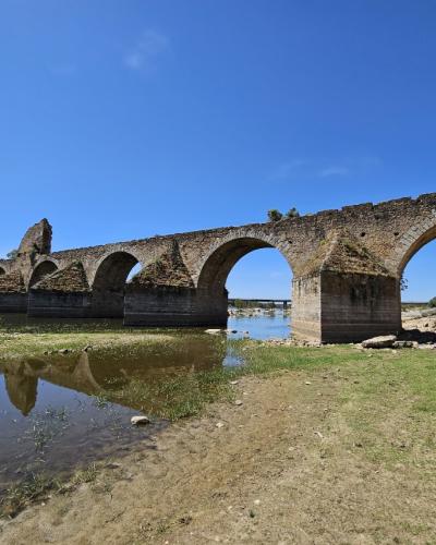 A long, multi-arched medieval stone bridge in a state of partial ruin, spanning a river with low water levels under a clear blue sky.