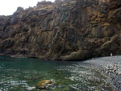 A rocky cove with a pebble beach and a steep cliff face featuring prominent columnar jointing in volcanic rock, with a person standing on the shore.