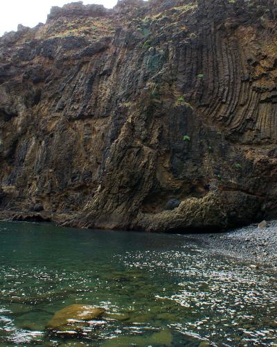 A rocky cove with a pebble beach and a steep cliff face featuring prominent columnar jointing in volcanic rock, with a person standing on the shore.