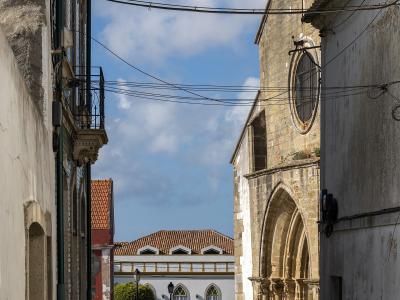 A narrow cobblestone street in a Portuguese village featuring a stone church with a Gothic-style arched portal and rose window on the right, and a white car parked in the distance.