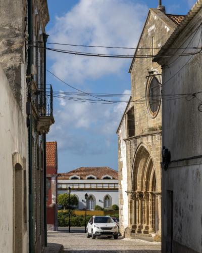 A narrow cobblestone street in a Portuguese village featuring a stone church with a Gothic-style arched portal and rose window on the right, and a white car parked in the distance.