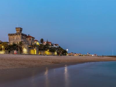 A long sandy beach at twilight with a large, illuminated stone castle-like building (Palácio do Freixo) on the left and a pier extending into the sea on the right.