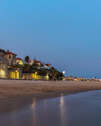 A long sandy beach at twilight with a large, illuminated stone castle-like building (Palácio do Freixo) on the left and a pier extending into the sea on the right.