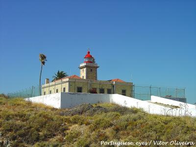 The image shows a stone lighthouse with a red lantern room and a red dome, situated on a grassy coastal hill with a white perimeter wall and a fence, under a clear blue sky.