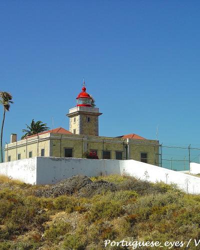 The image shows a stone lighthouse with a red lantern room and a red dome, situated on a grassy coastal hill with a white perimeter wall and a fence, under a clear blue sky.