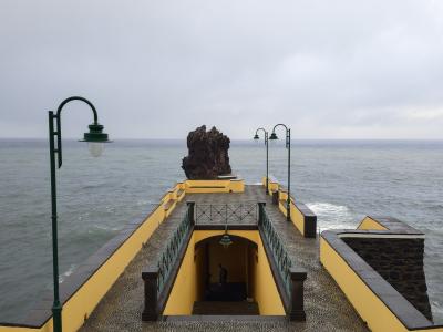 A yellow and black stone pier extending into the ocean, featuring a distinctive large rock formation at its end and green lampposts along the walkway.