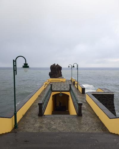A yellow and black stone pier extending into the ocean, featuring a distinctive large rock formation at its end and green lampposts along the walkway.