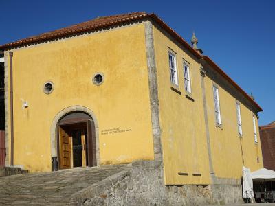 A yellow colonial-style building with a tiled roof, stone foundation, and arched entrance, featuring text on the facade that reads "ALFÂNDEGA RÉGIA MUSEU DA CONSTRUÇÃO NAVAL".
