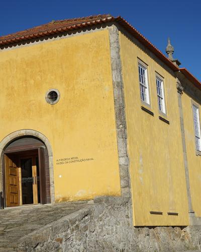 A yellow colonial-style building with a tiled roof, stone foundation, and arched entrance, featuring text on the facade that reads "ALFÂNDEGA RÉGIA MUSEU DA CONSTRUÇÃO NAVAL".