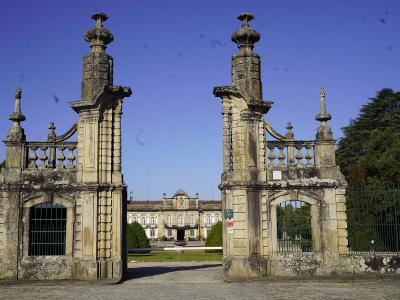 A view through a grand, ornate stone gate with tall pillars and balustrades, looking towards a large neoclassical palace building in the distance across a lawn.