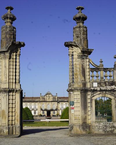 A view through a grand, ornate stone gate with tall pillars and balustrades, looking towards a large neoclassical palace building in the distance across a lawn.