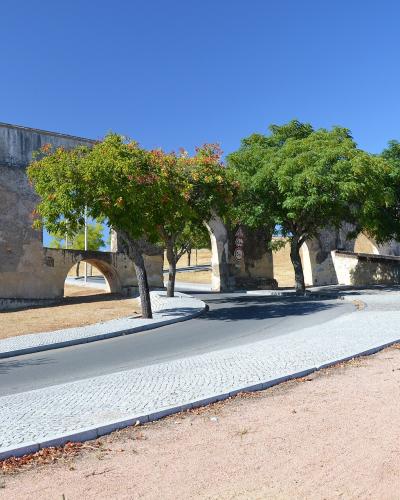A low-angle view of a long, multi-tiered stone aqueduct with rounded arches running parallel to a paved road lined with trees under a clear blue sky.