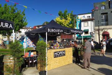 An outdoor craft beer festival stall with a black canopy labeled "BURGUESA" and "FERMENTAGE," a wooden counter with a sign reading "HOPEN CRAFT BEER FESTIVAL," people serving and attending, colorful bunting overhead, and buildings in the backgroun