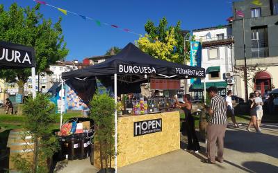 An outdoor craft beer festival stall with a black canopy labeled "BURGUESA" and "FERMENTAGE," a wooden counter with a sign reading "HOPEN CRAFT BEER FESTIVAL," people serving and attending, colorful bunting overhead, and buildings in the backgroun