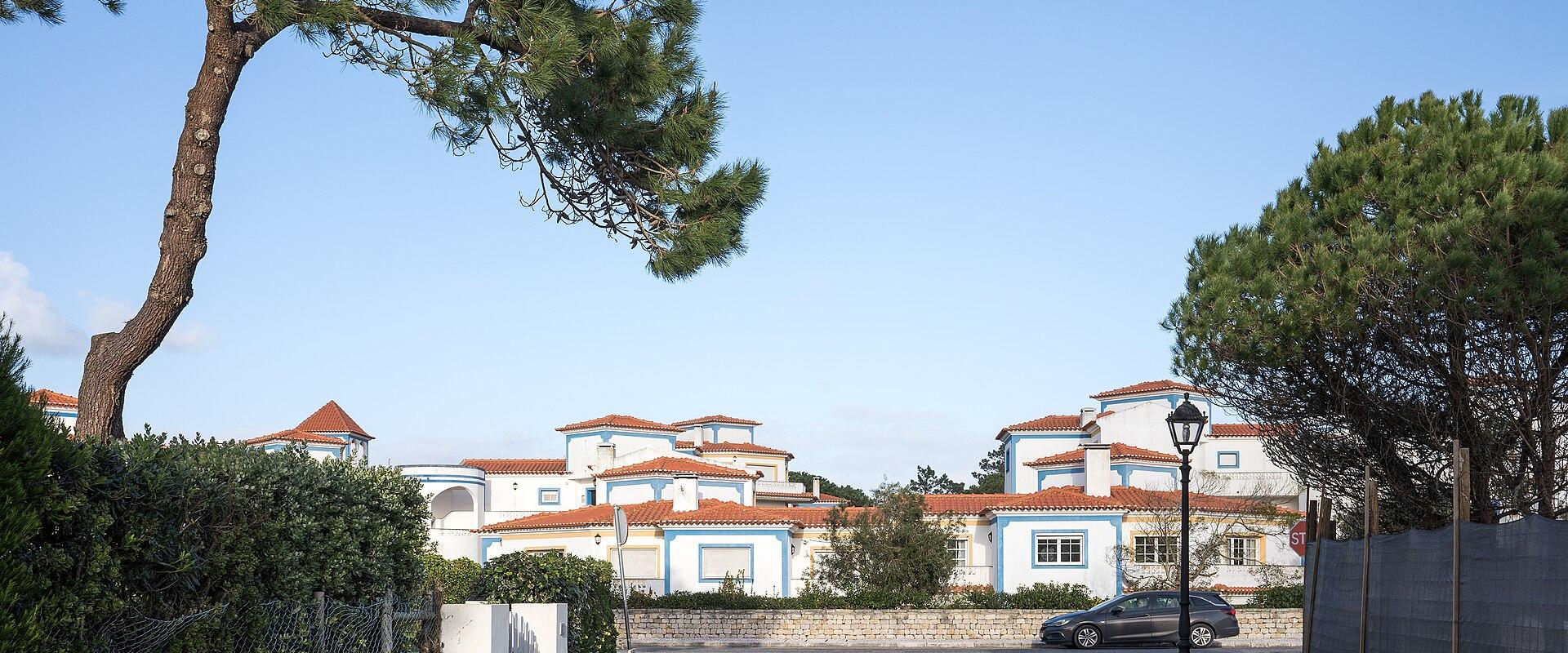 A paved road curves past white villas with blue trim and terracotta roofs, framed by pine trees and a stone wall under a clear sky.