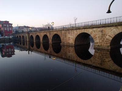 A long, multi-arched Roman stone bridge spanning a calm river at dusk, with a red building and street lamps visible on the bank.