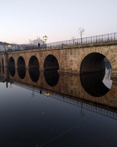 A long, multi-arched Roman stone bridge spanning a calm river at dusk, with a red building and street lamps visible on the bank.