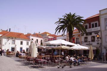 An outdoor plaza with cobblestone paving, white buildings with terracotta roofs, and a large palm tree, featuring an outdoor cafe with white umbrellas and a statue of a figure in a cloak.