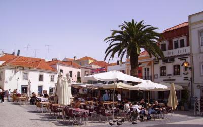 An outdoor plaza with cobblestone paving, white buildings with terracotta roofs, and a large palm tree, featuring an outdoor cafe with white umbrellas and a statue of a figure in a cloak.