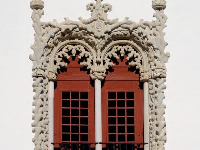 A close-up architectural detail of an ornate Manueline-style window frame with red shutters and a black iron railing against a white wall.