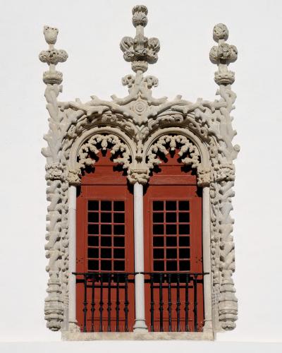 A close-up architectural detail of an ornate Manueline-style window frame with red shutters and a black iron railing against a white wall.