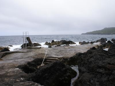 A coastal swimming area built into black volcanic rock, featuring a concrete diving platform with a metal ladder, steps leading into the water, and the Atlantic Ocean in the background under an overcast sky.