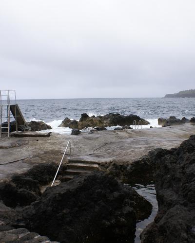 A coastal swimming area built into black volcanic rock, featuring a concrete diving platform with a metal ladder, steps leading into the water, and the Atlantic Ocean in the background under an overcast sky.