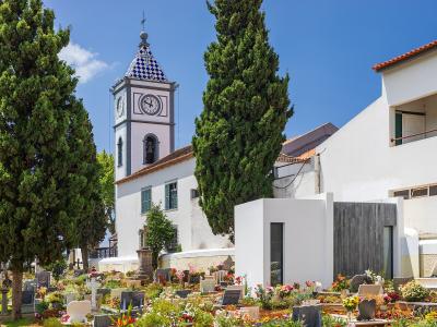 A white church with a distinctive blue and white checkered tile clock tower stands behind a cemetery filled with headstones and flowers under a clear blue sky.
