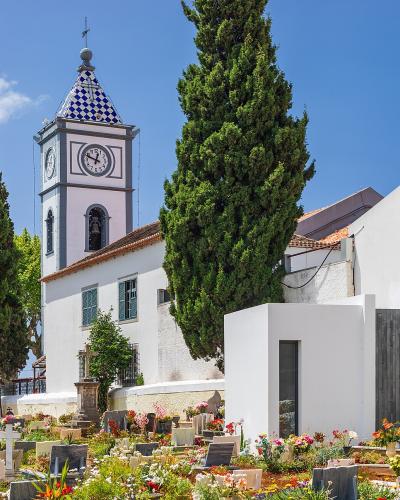 A white church with a distinctive blue and white checkered tile clock tower stands behind a cemetery filled with headstones and flowers under a clear blue sky.