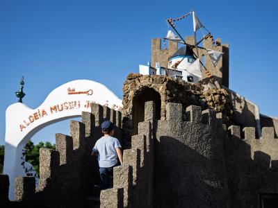 A child in a blue shirt stands on the battlements of a rustic stone castle structure, with a white archway to the left bearing the text "ALDEIA MUSEU JOSÉ FRANCO".