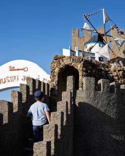 A child in a blue shirt stands on the battlements of a rustic stone castle structure, with a white archway to the left bearing the text "ALDEIA MUSEU JOSÉ FRANCO".