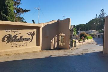 The image shows the entrance gate and signage for the "blue & green Vilalara THALASSA RESORT," a five-star establishment, set against a clear blue sky with surrounding vegetation.