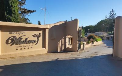 The image shows the entrance gate and signage for the "blue & green Vilalara THALASSA RESORT," a five-star establishment, set against a clear blue sky with surrounding vegetation.