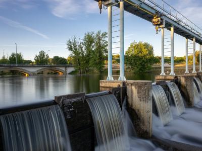 A long-exposure photograph of a concrete weir or small dam on a river, with water cascading over spillways, a metal gantry crane structure overhead, and an arched bridge visible in the background across the river.
