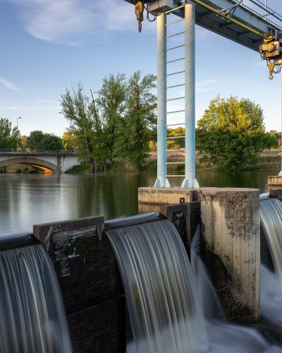 A long-exposure photograph of a concrete weir or small dam on a river, with water cascading over spillways, a metal gantry crane structure overhead, and an arched bridge visible in the background across the river.