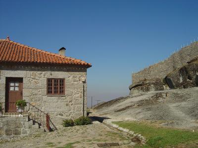 A stone building with a terracotta tiled roof and a wooden door stands in the foreground next to a large, curved stone fortification wall built upon a rocky outcrop under a clear blue sky.