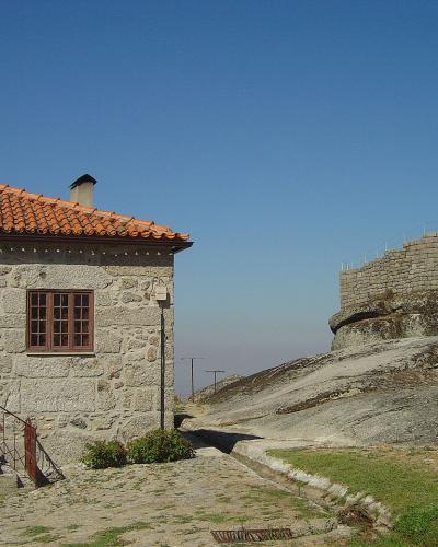 A stone building with a terracotta tiled roof and a wooden door stands in the foreground next to a large, curved stone fortification wall built upon a rocky outcrop under a clear blue sky.