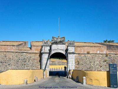 A stone archway gateway set within a fortified wall featuring yellow-painted lower sections and a stone crest above the arch, with a blue information sign to the right.