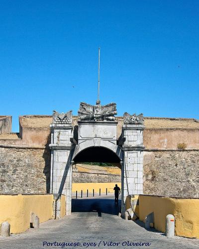 A stone archway gateway set within a fortified wall featuring yellow-painted lower sections and a stone crest above the arch, with a blue information sign to the right.