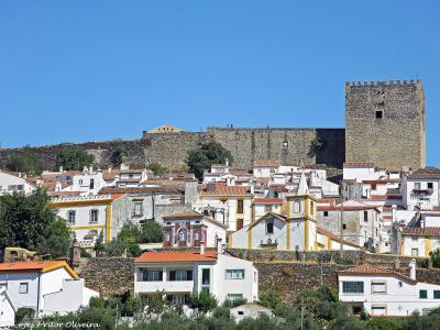 A view of a hilltop town with white houses and terracotta roofs, dominated by a large medieval stone castle with a prominent square keep and crenellated walls on the ridge above.