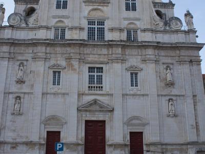 A low-angle photograph of the ornate Baroque facade of a large stone church with a central cross, statues in niches, and three red doors at the base, with cars parked on a cobblestone street in front.
