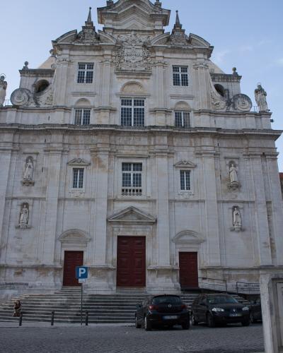 A low-angle photograph of the ornate Baroque facade of a large stone church with a central cross, statues in niches, and three red doors at the base, with cars parked on a cobblestone street in front.
