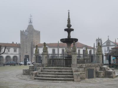 A stone fountain with a multi-tiered basin and decorative finial stands in the center of a paved public square, surrounded by a low iron fence and stone posts, with a historic stone tower and white buildings in the background under an overcast sky.