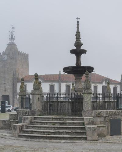 A stone fountain with a multi-tiered basin and decorative finial stands in the center of a paved public square, surrounded by a low iron fence and stone posts, with a historic stone tower and white buildings in the background under an overcast sky.