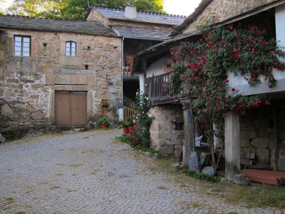A cobblestone street in a traditional stone village, featuring a building with a large wooden door and another structure with a balcony covered in red flowering vines.