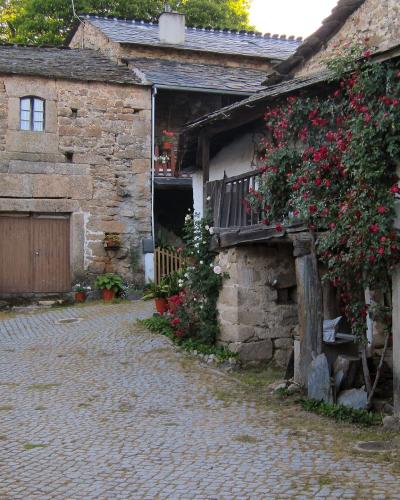 A cobblestone street in a traditional stone village, featuring a building with a large wooden door and another structure with a balcony covered in red flowering vines.