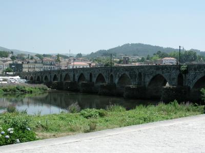 A long, multi-arched stone bridge spanning a wide river, with a town and a church tower visible in the background under a clear sky.