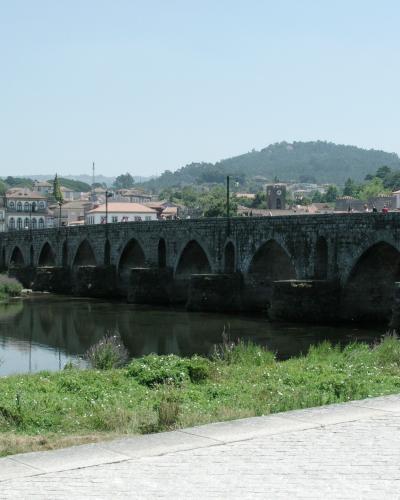A long, multi-arched stone bridge spanning a wide river, with a town and a church tower visible in the background under a clear sky.