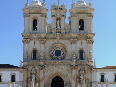 A full frontal view of the ornate, light-colored stone facade of a large church or monastery featuring twin bell towers, a central rose window, and a grand arched entrance under a clear blue sky.