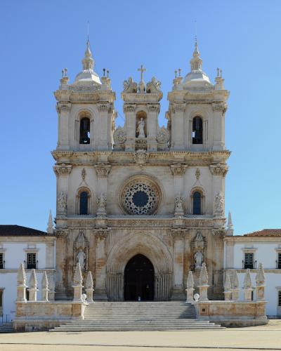 A full frontal view of the ornate, light-colored stone facade of a large church or monastery featuring twin bell towers, a central rose window, and a grand arched entrance under a clear blue sky.